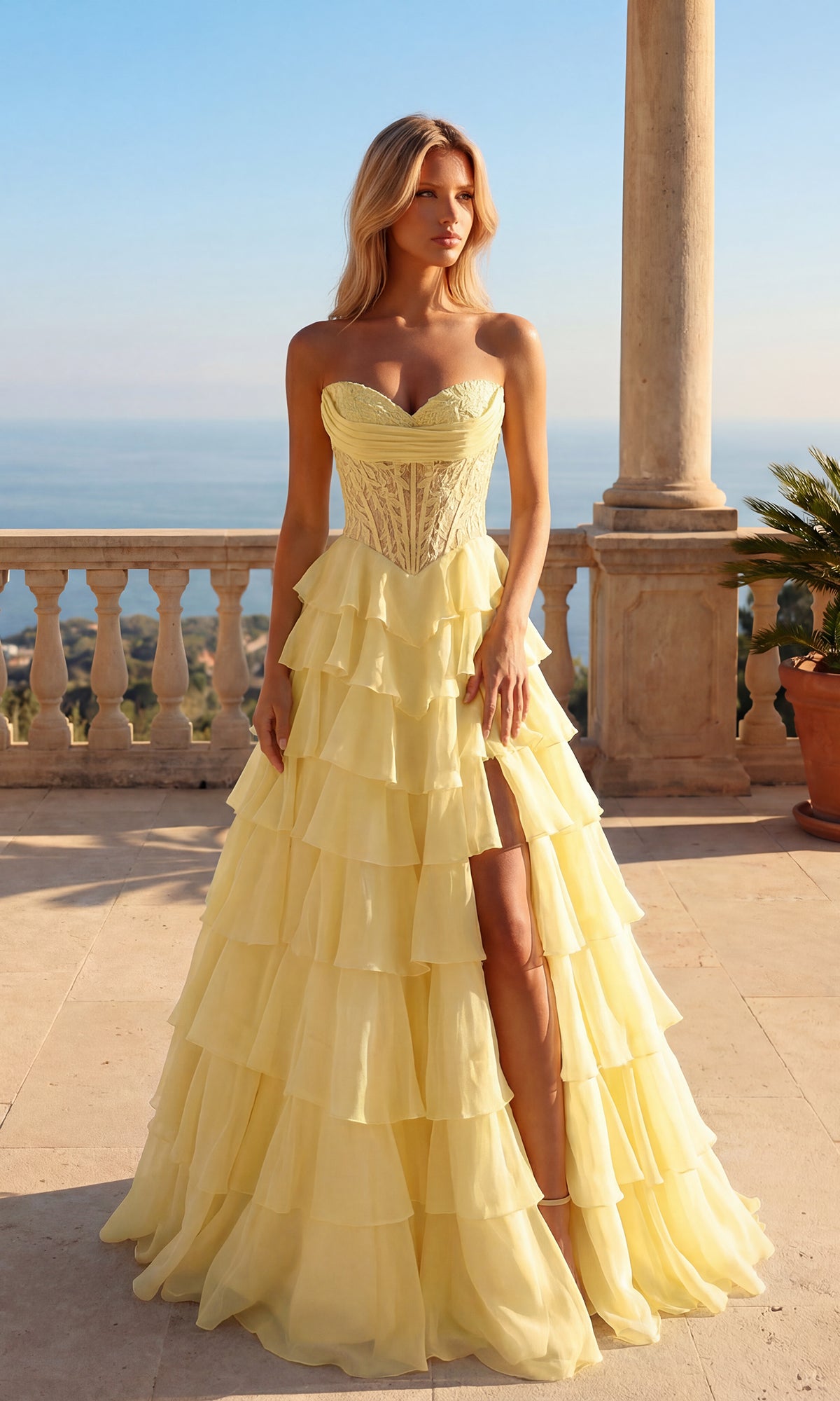 A woman wears the Long Ruffle Prom Gown: PromGirl Exclusive 250205 in light yellow, featuring a strapless sweetheart bodice and ruffled skirt, as she stands on a stone terrace overlooking water and greenery beneath a clear sky.